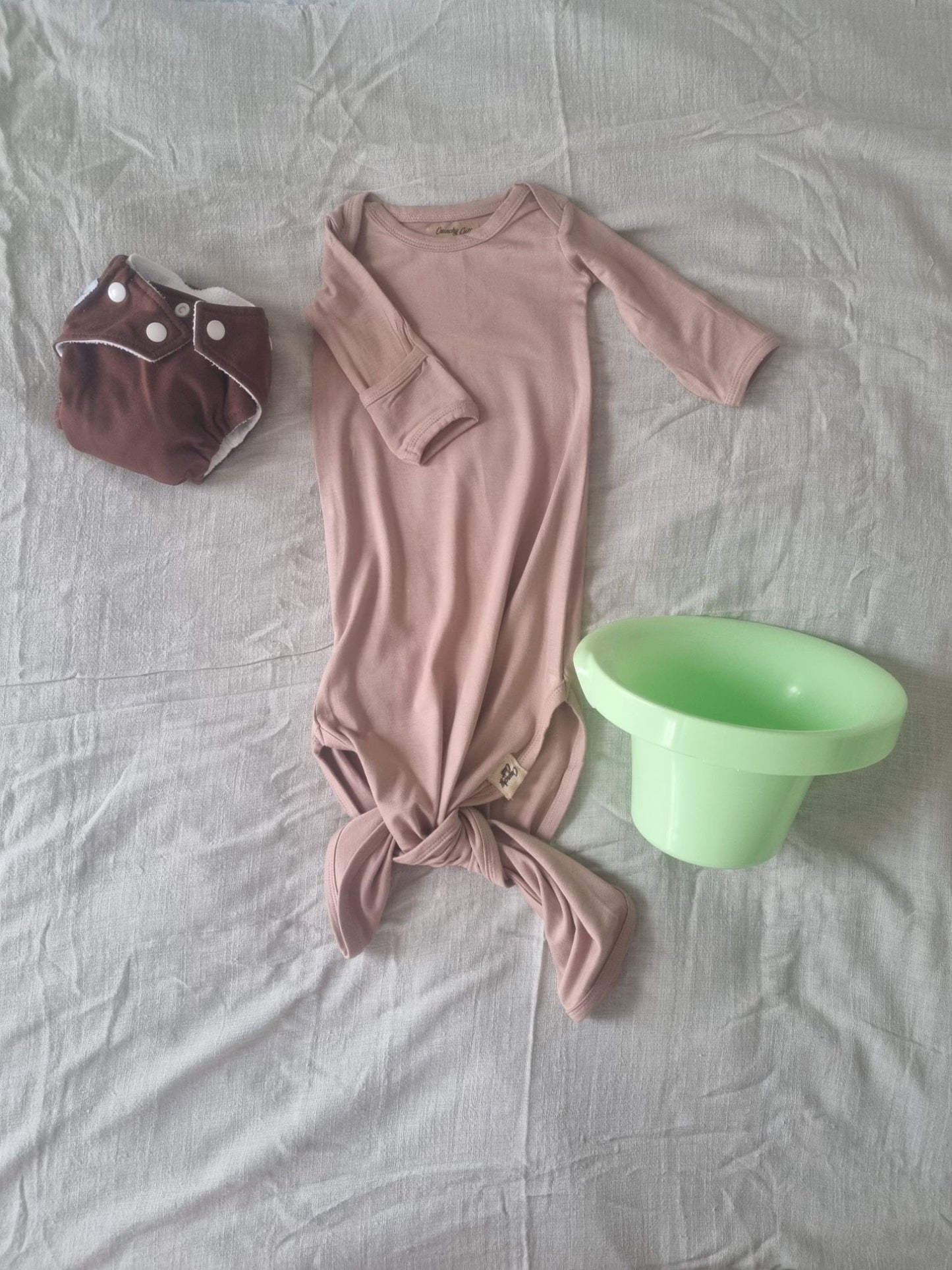 Brown knotted baby gown, front view, rolled rolled sleve, decorated background with grey linen sheet and top hat potty and grey cloth nappy.