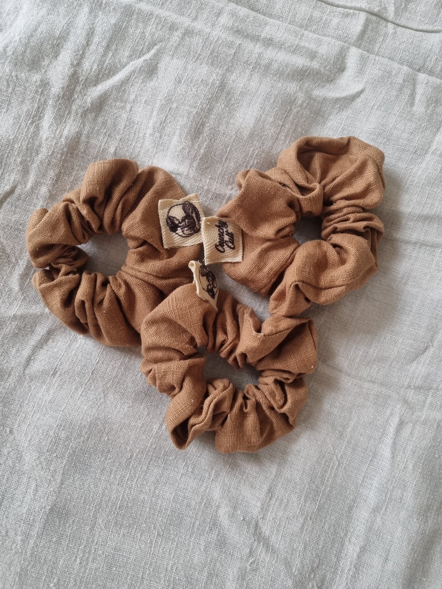 Three linen brown hair scrunches modelled on a grey linen sheet
