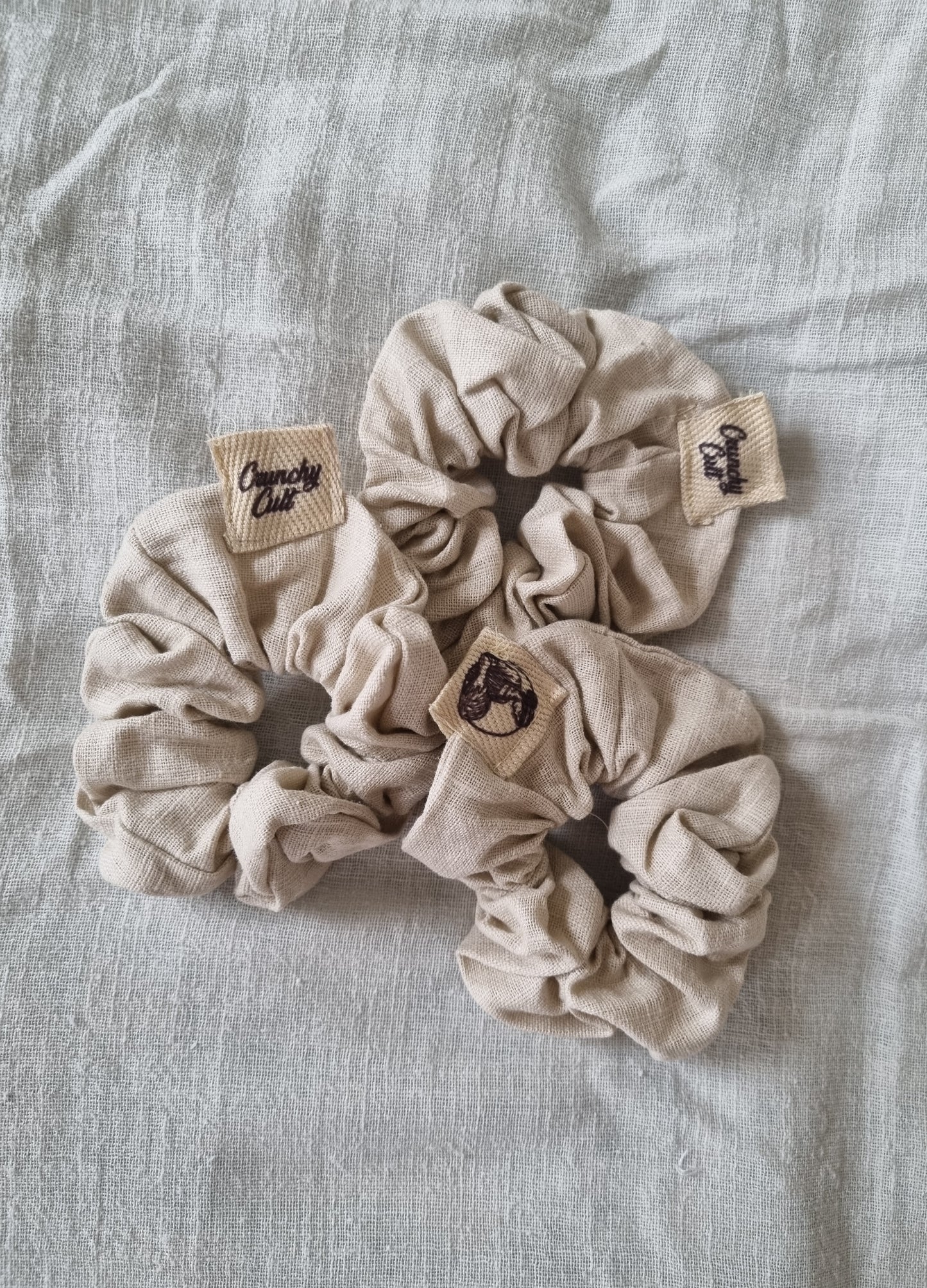 Three linen baige hair scrunches modelled on a grey linen sheet