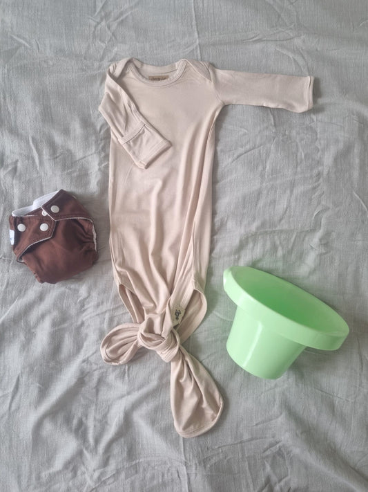 Baige knotted baby gown, front view, rolled rolled sleve, decorated background with grey linen sheet, top hat potty and brown cloth nappy.