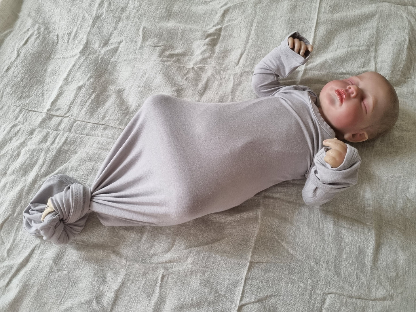 Grey knotted baby gown modeled by baby on grey linen sheet. Tilted top side view