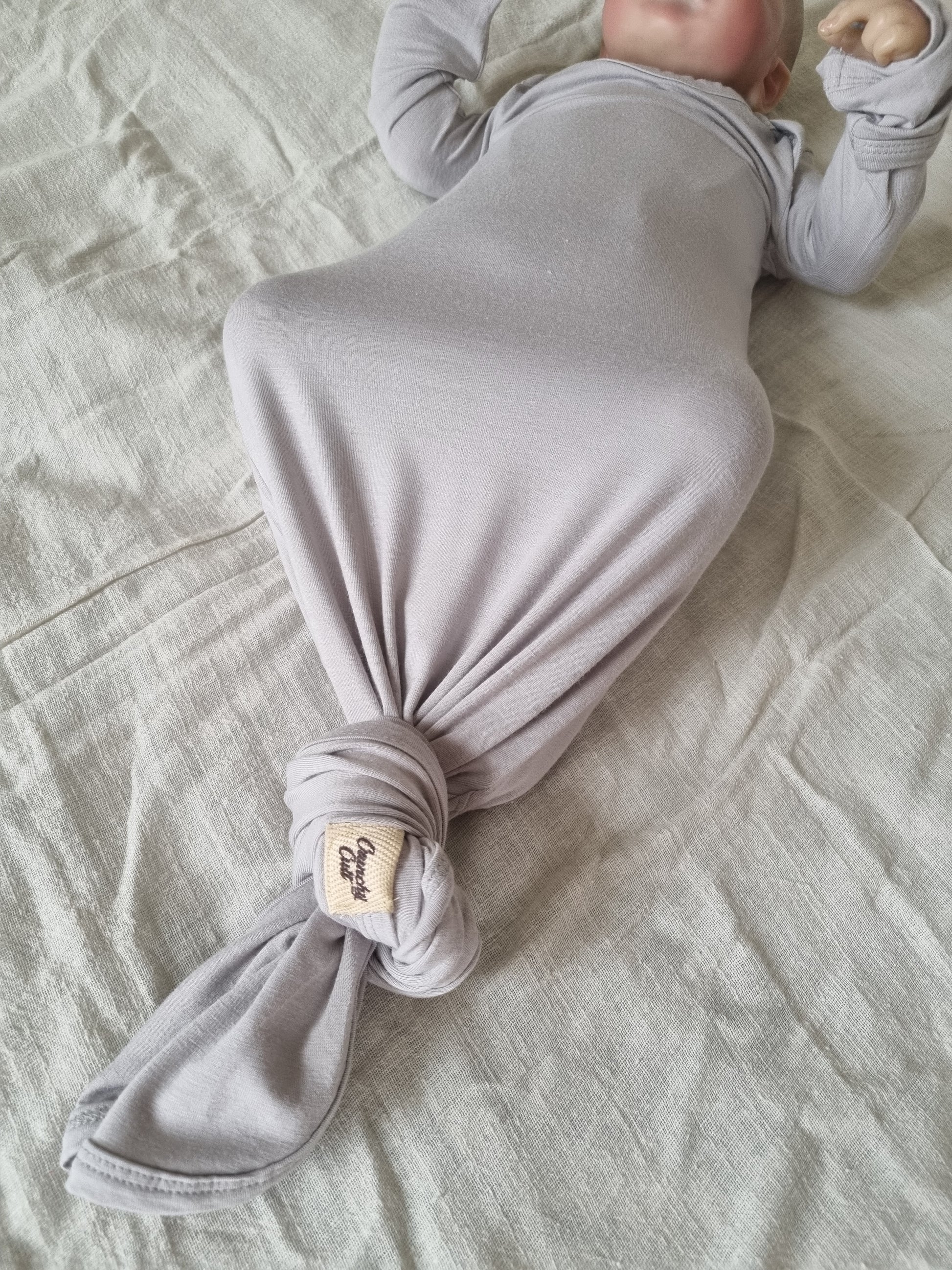 Grey knotted baby gown, modeled by baby showing the bottom view with a knot and logo
